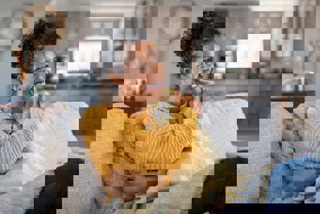 A smiling woman is wrapped in a blanket and sits on a gray sofa, looking content