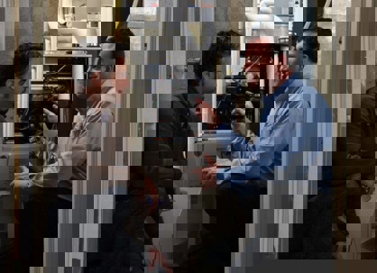 Two men are inspecting a furnace in a utility closet.