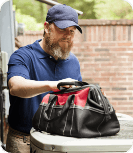 A bearded man wearing a blue uniform and cap is servicing an HVAC unit