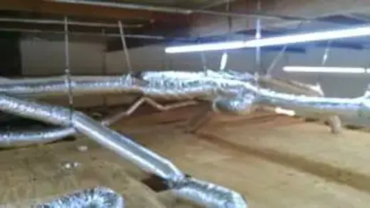 A cluttered crawl space with ductwork and fluorescent lights